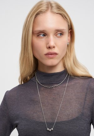 Young woman with blonde hair wearing a sheer dark gray turtleneck and layered silver necklaces, looking to the right against a white background.
