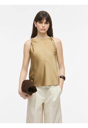 Femme portant un haut beige sans manches et un pantalon blanc, tenant une veste marron pliée, avec des cheveux raides foncés et une expression neutre.