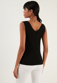 Black sleeveless top with a V-neck back and smooth texture. Features a decorative chain accent at the neckline. Worn with white pants.