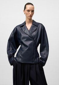 Donna che indossa una giacca di pelle blu scuro con ampi risvolti e chiusura con zip, abbinata a pantaloni neri larghi, in piedi su uno sfondo uniforme.