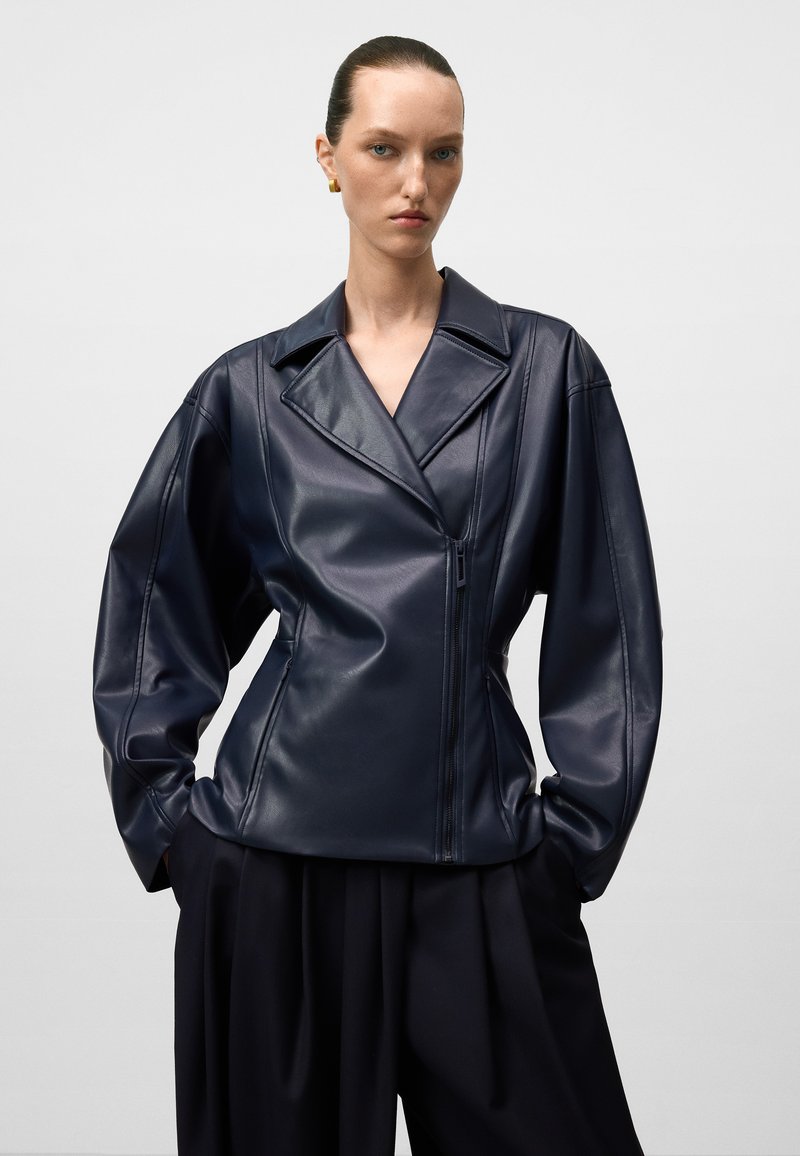 Donna che indossa una giacca di pelle blu scuro con ampi risvolti e chiusura con zip, abbinata a pantaloni neri larghi, in piedi su uno sfondo uniforme.