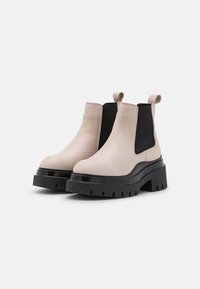 Bottines en cuir beige avec un panneau latéral élastique noir, semelle noire épaisse et tirette pour un enfilage facile. Semelle plate et texturée.