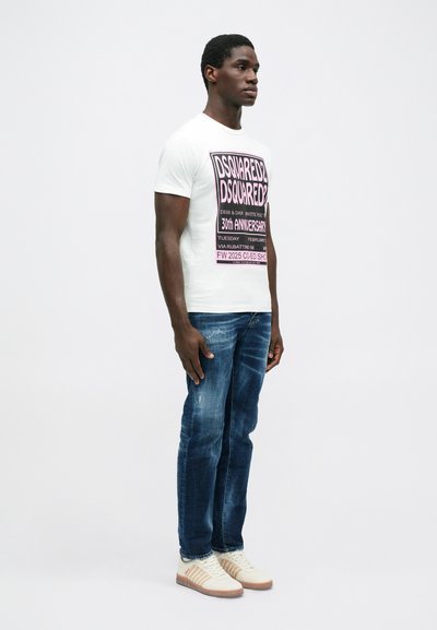 Weißes T-Shirt mit pinkem und schwarzem grafischem Text, blaue Jeans und beige Sneaker mit Streifen. Das Model steht mit entspannten Armen.