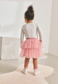 Gestufter pinker Tüllrock mit elastischem Bund, getragen über einem hellgrauen Langarmoberteil; weiße Sneakers mit pinken Akzenten und Herzdetail.