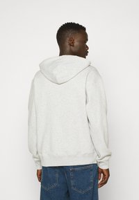 Pilkas gobtuvas megztinis, pagamintas iš švelnaus medvilnės mišinio, su laisvu kirpimu ir gumuotomis rankovėmis, derinamas su mėlynomis džinsinėmis kelnėmis.