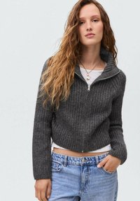 Siva rebrasta skrajšana zip-up pulover z dolgimi rokavi, v kombinaciji s svetlo modrimi kavbojkami. Tekstura se zdi mehka in prijetna, primerna za sproščeno nošenje.