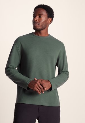 Green textured long-sleeve shirt with a round neckline, featuring a relaxed fit and ribbed pattern, paired with dark pants.