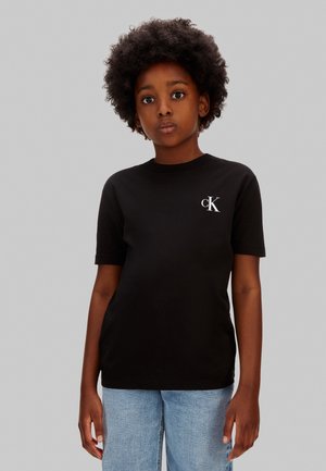 Tânăr cu păr creț, purtând un tricou negru Calvin Klein și blugi albastru deschis, stând în fața unui fundal gri simplu.