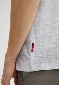 Weißes, strukturiertes T-Shirt mit einem roten "Joopi Jeans"-Label an der linken Seitennaht, getragen mit grauen gemusterten Hosen.