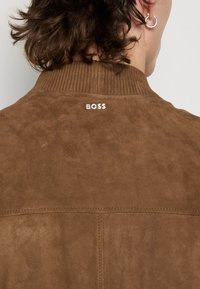 Veste en suède marron avec un col côtelé, présentant des lettres blanches "BOSS" au dos. La texture semble douce, avec des détails de couture visibles.