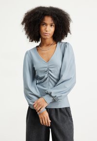 Maglietta azzurra chiara a maniche lunghe con scollatura a V, con dettagli arricciati sul davanti e maniche a sbuffo. Tessuto morbido e vestibilità rilassata.