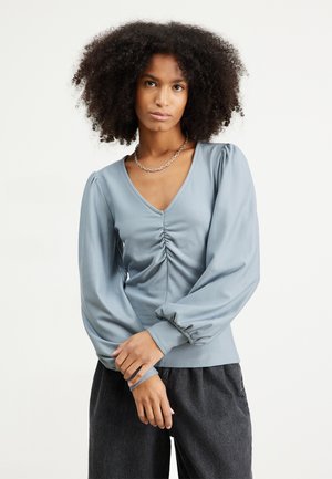 Light blue long-sleeve top with a V-neck, featuring ruched detailing at the front and puffed sleeves. Soft fabric and relaxed fit.