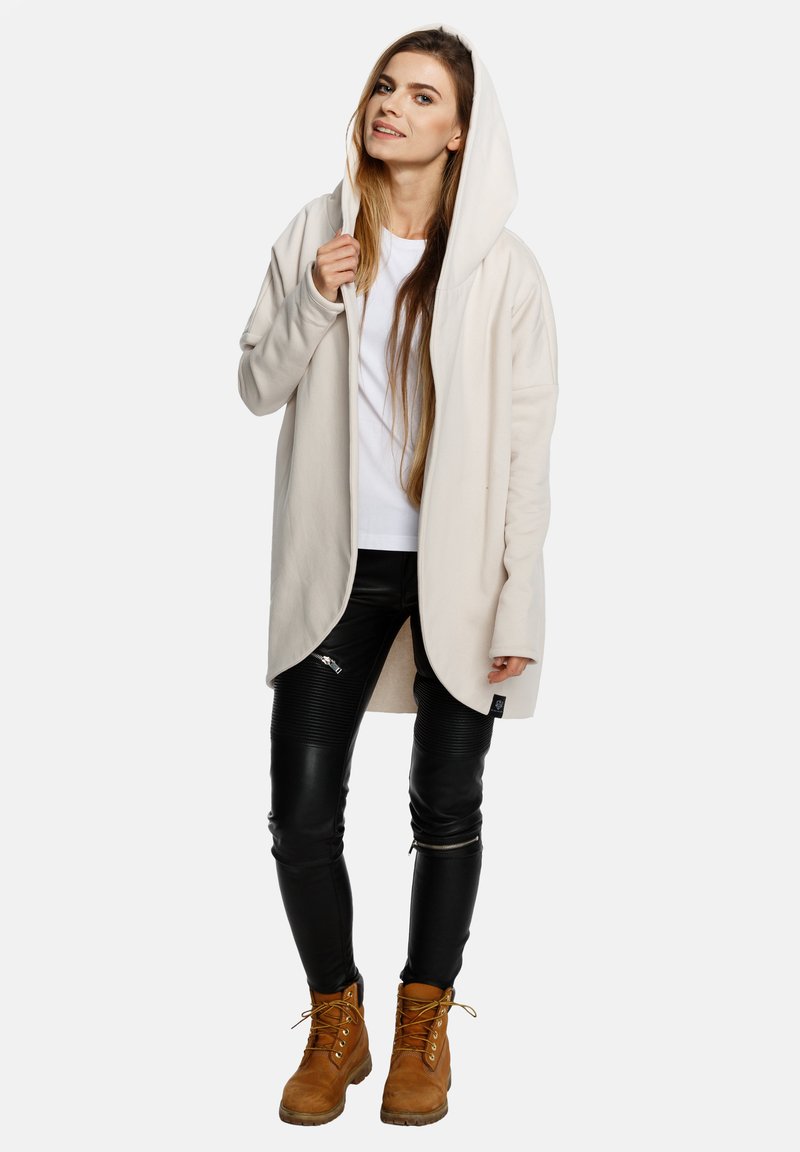 Jasno-beżowa kurtka z kapturem o gładkiej fakturze, zestawiona z czarnymi skórzanymi spodniami z karbowanymi akcentami oraz brązowymi butami na sznurowadłach.