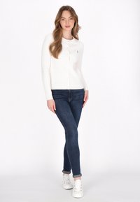 Witte geribde cardigan met knopen aan de voorkant en lange mouwen, gecombineerd met donkerblauwe skinny jeans en witte sneakers. Strakke lijnen, casual outfit.