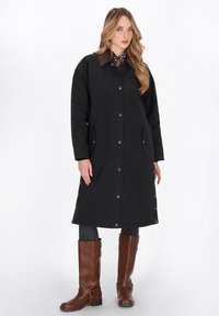 Kvinde iført en lang sort knapcoat med en brun krave, mørke jeans, brune støvler til knæene og et mønstret tørklæde, stående på hvid baggrund.