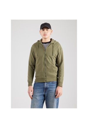 Olivengroene hoodie met ritssluiting, voorzien van een capuchon, frontzakken en geribbelde manchetten, gedragen over een zwart-wit gestreept shirt, gecombineerd met een blauwe spijkerbroek.