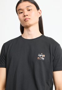 Tricou negru din bumbac cu un mic logo multicolor "Alpha Industries" pe piept. Mâneci scurte și guler rotund.