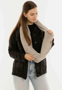 Jonge vrouw met lang bruin haar, gekleed in een zwarte spijkerjacket en lichte jeans, houdt een grijze gebreide infinity sjaal vast en kijkt ernaar.