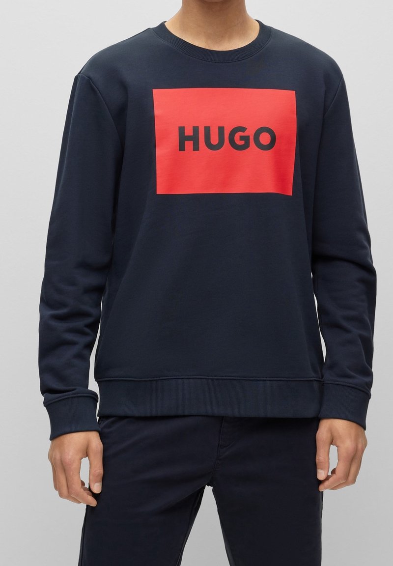 Sudadera azul marino de tejido suave, presenta un gráfico rectangular rojo con "HUGO" en texto negro en la parte delantera. Puños y dobladillo acanalados.