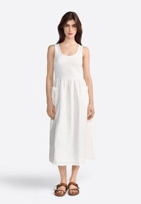 Vestido de lino blanco con un top ajustado estilo tank y una falda acampanada. Presenta dos bolsillos frontales y se lleva con sandalias marrones de tiras.