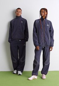 Két ember, akik azonos sötét Adidas melegítőfelsőt viselnek, egy fehér fal előtt állnak egy zöld padlón, előrefelé nézve, semleges arccal.