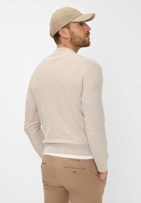 Beige gestrickter Pullover mit rundem Halsausschnitt und geripptem Saum, getragen über hellen Hosen. Eine passende beige Mütze vervollständigt den Look.