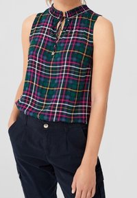 Top senza maniche a quadri verdi con dettaglio al collo a fiocco, indossato con pantaloni blu navy a vita alta, mani casualmente nelle tasche.
