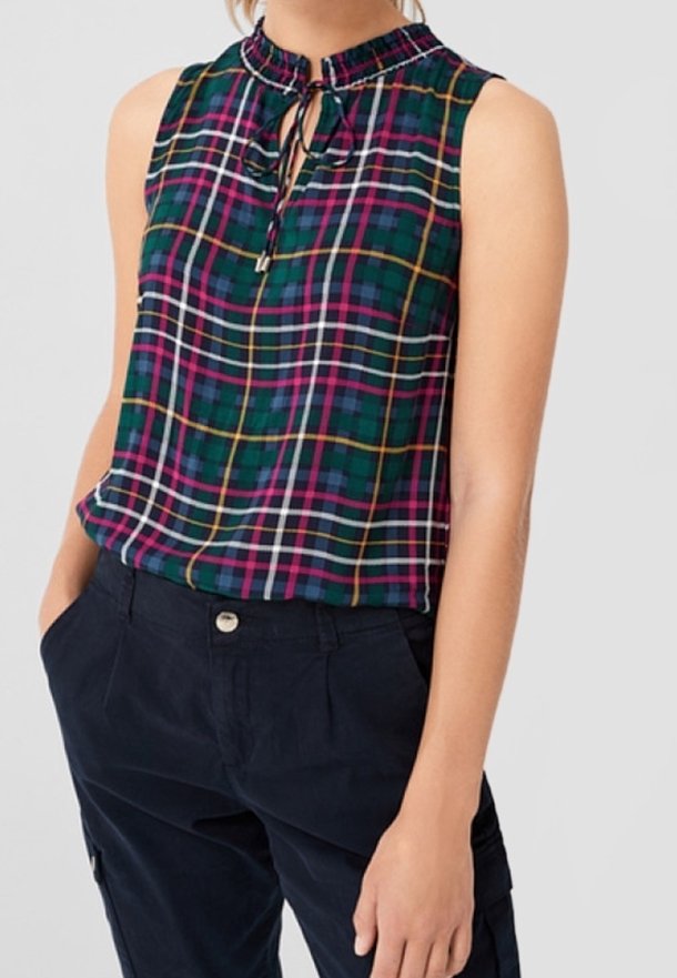 Top senza maniche a quadri verdi con dettaglio al collo a fiocco, indossato con pantaloni blu navy a vita alta, mani casualmente nelle tasche.