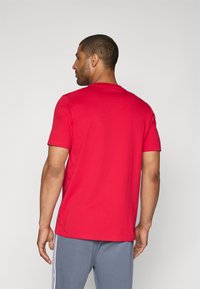 Rød bomulds T-shirt med korte ærmer og rund halsudskæring, set bagfra. Sammen med grå træningsbukser med sidelæg.