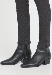 Black leather ankle boots with a pointed toe and a low block heel. Textured upper with seamless design. Paired with patterned black pants.