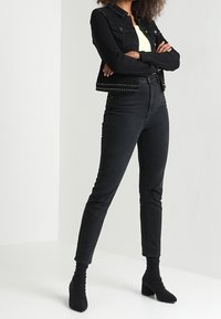 Chaqueta de denim negra con detalles de tachuelas, combinada con jeans negros de cintura alta y botines negros de punta. La modelo se encuentra con los brazos cruzados.