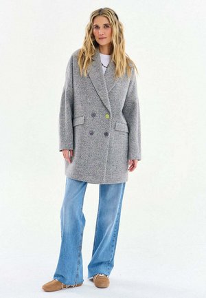 Kobieta o długich blond włosach, ubrana w szary dwurzędowy płaszcz w jodełkę, jasnoniebieskie szerokie spodnie dżinsowe i brązowe buty, stoi na białym tle.