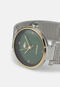 Reloj Lacoste redondo con esfera verde, acentos dorados, correa de malla plateada y marcadores de cristal. Presenta un detalle de corona verde.