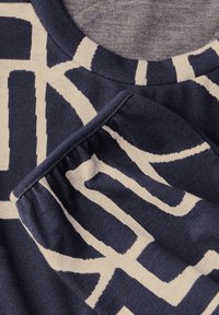 Gros plan d'un tissu bleu marine avec un motif géométrique crème abstrait et un col côtelé détaillé ainsi qu'un poignet de manches froncé.