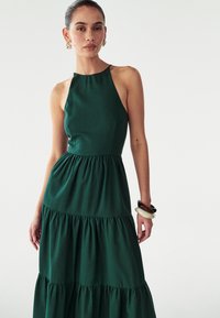 Zelena, brez rokavov maksi obleka s prilegajočim se zgornjim delom in večplastnim, nabranim krilom. Gladka tekstura blaga, minimalističen dizajn, z visokim ovratnikom.