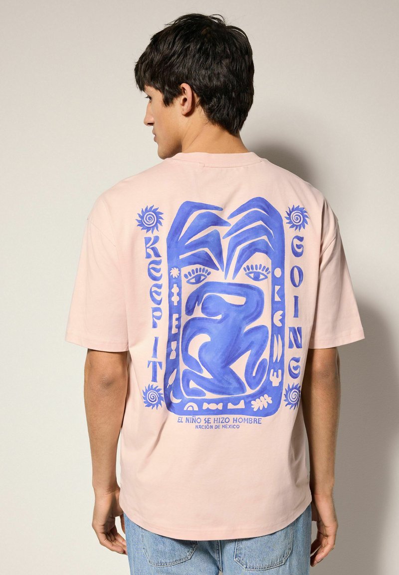 Camiseta rosa claro con un gran diseño gráfico en azul de manos y una figura en la parte trasera. Manga corta, corte relajado y material de algodón.