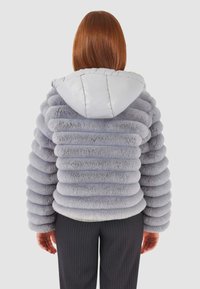 Giacca in finta pelliccia grigia con design arrotondato e a coste e cappuccio grigio chiaro. Presenta un orlo inferiore elasticizzato e una texture morbida. Indossata con pantaloni a righe.
