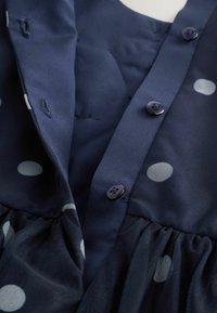 Robe bleu marine à pois blancs, avec un devant boutonné, une jupe plissée et une texture lisse. Les boutons sont ronds et foncés.