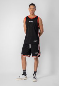 Camiseta sin mangas de baloncesto de malla negra con ribete rojo, combinada con pantalones cortos negros con franjas rojas y blancas. Lleva puesta unas zapatillas blancas.
