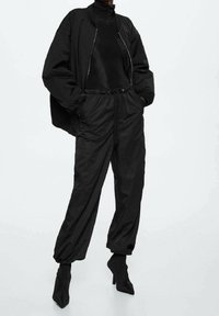 Zwarte bomberjack met ritssluiting, gedragen over een zwarte coltrui, gecombineerd met loszittende zwarte broek met elastische manchetten en een strikband in de taille.