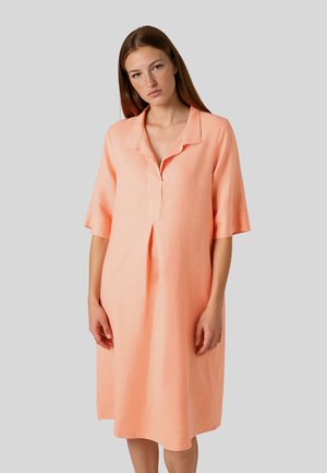 Junge Frau mit glattem braunem Haar, die ein lockeres pfirsichfarbenes knielanges Kleid mit kurzen Ärmeln und kleinem Kragen trägt.