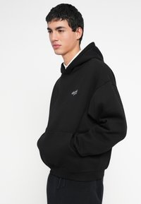 Fiatal férfi fekete kapucnis pulóverben fehér logóval, kezével a zsebében, kissé balra nézve, sötét, texturált nadrágot viselve, egyszerű háttér előtt.