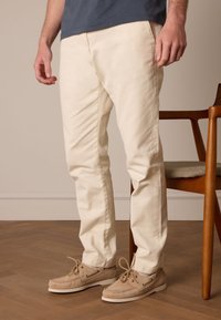 Helle beige Baumwollhosen mit schlankem Schnitt, die eine subtile Textur aufweisen. Kombiniert mit beigen Bootsschuhen und steht neben einem Holzhocker.