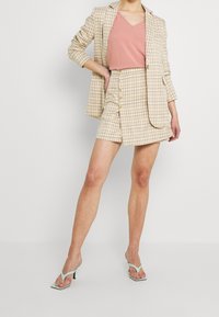 Beżowa kratkowana marynarka o dopasowanym kroju, zestawiona z różowym topem z dekoltem w serek i beżową spódnicą na guziki. Stylizację dopełniają zielone sandały na obcasie.