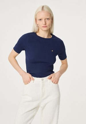 Vrouw met steil blond haar draagt een marineblauw T-shirt met korte mouwen en witte, gestructureerde, hooggetailleerde broek, staat met handen in de zakken.
