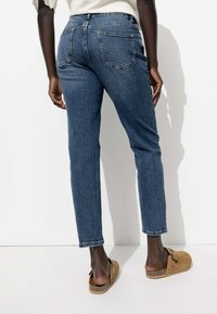 Jean en denim bleu avec une coupe ajustée, taille mi-haute et deux poches arrière. Associé à des sabots marron à enfiler sur un fond uni.
