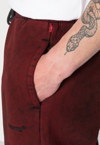 Calças de corduroy bordeaux com bolsos laterais, apresentando uma etiqueta vermelha da Levi's e um logo bordado. Textura suave e design de corte descontraído.