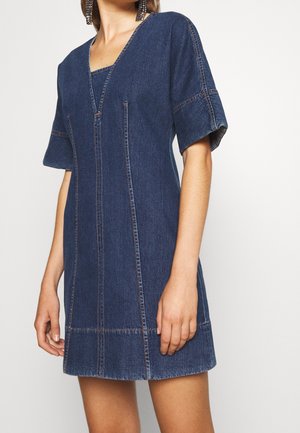 Vrouw draagt een donkerblauwe denimjurk met korte mouwen, een V-halslijn en zichtbare verticale en horizontale stiksel-details.