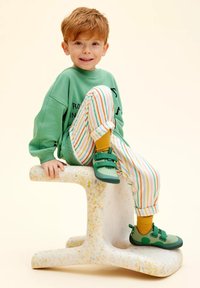 Groene sweatshirt met zwarte tekst, gestreepte broek in oranje en groen, gele sokken en groene schoenen. Pose op een gespikkelde, witte kruk.
