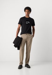Black cotton t-shirt with white Coca-Cola logo, beige trousers, and black shoes. Model holds a black jacket, showcasing a relaxed fit.